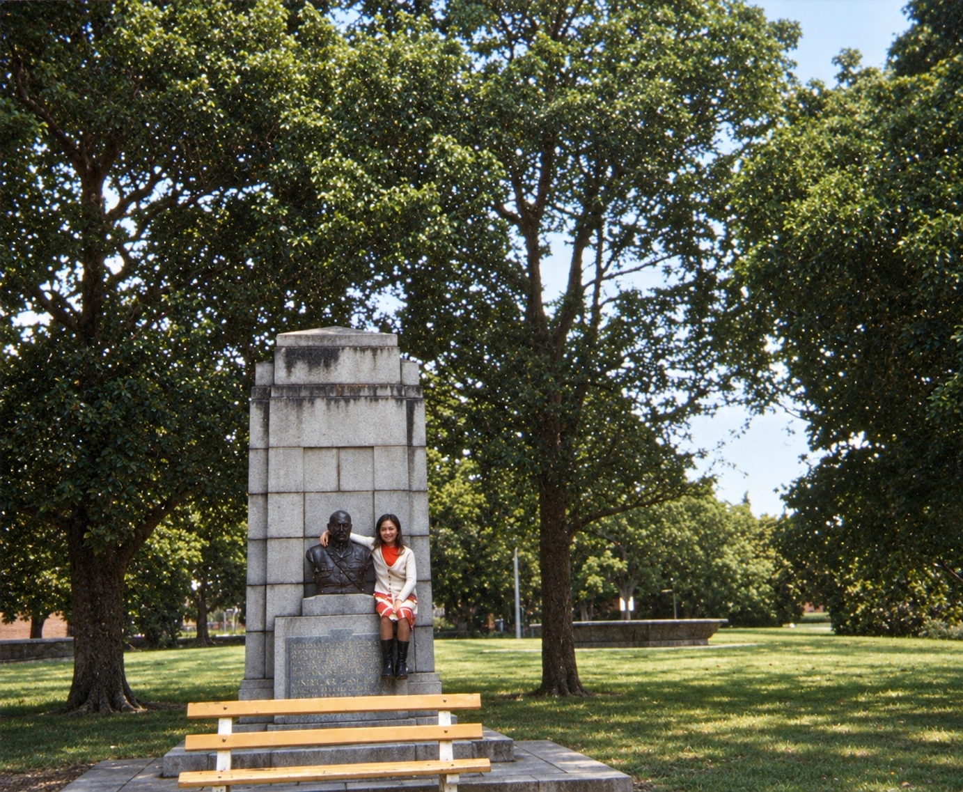 Restored colour photograph of a woman seated beside the bronze Monash bust on its stone memorial, with a yellow bench in front and large trees and lawn behind.