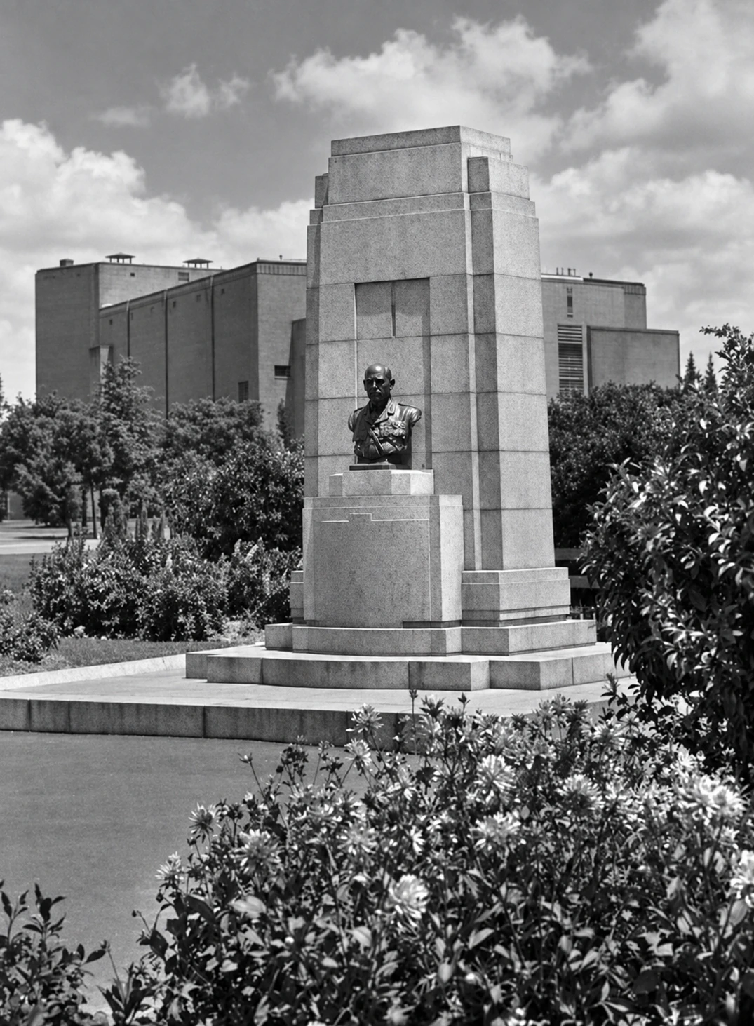 Restored black-and-white portrait view of the Sir John Monash bust and stepped stone memorial in landscaped gardens, with Yallourn Theatre behind it.