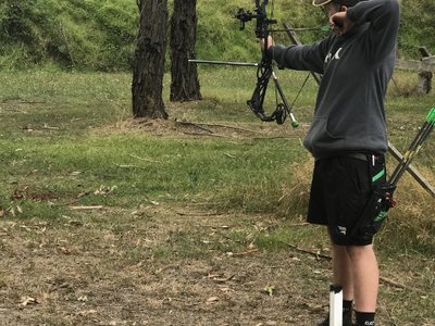 An archer taking aim at a 3D indoor target inside a clubroom, with colourful archery targets visible in the background.