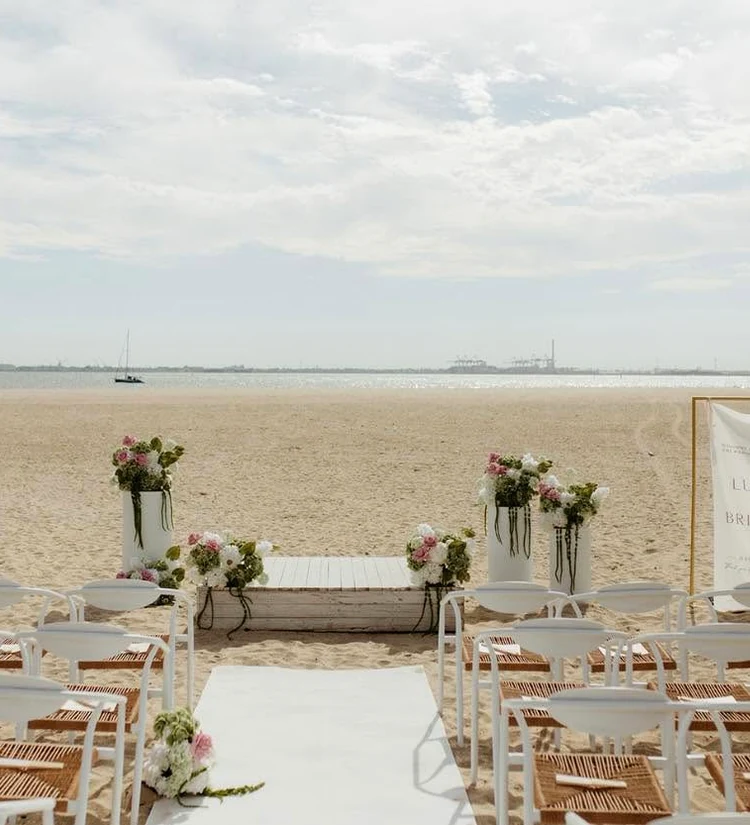 An outdoor beach wedding ceremony is arranged with pale floral installations around the aisle and platform.