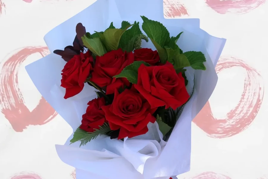 A bouquet of red roses wrapped in white paper against a pale pink painted-heart background.
