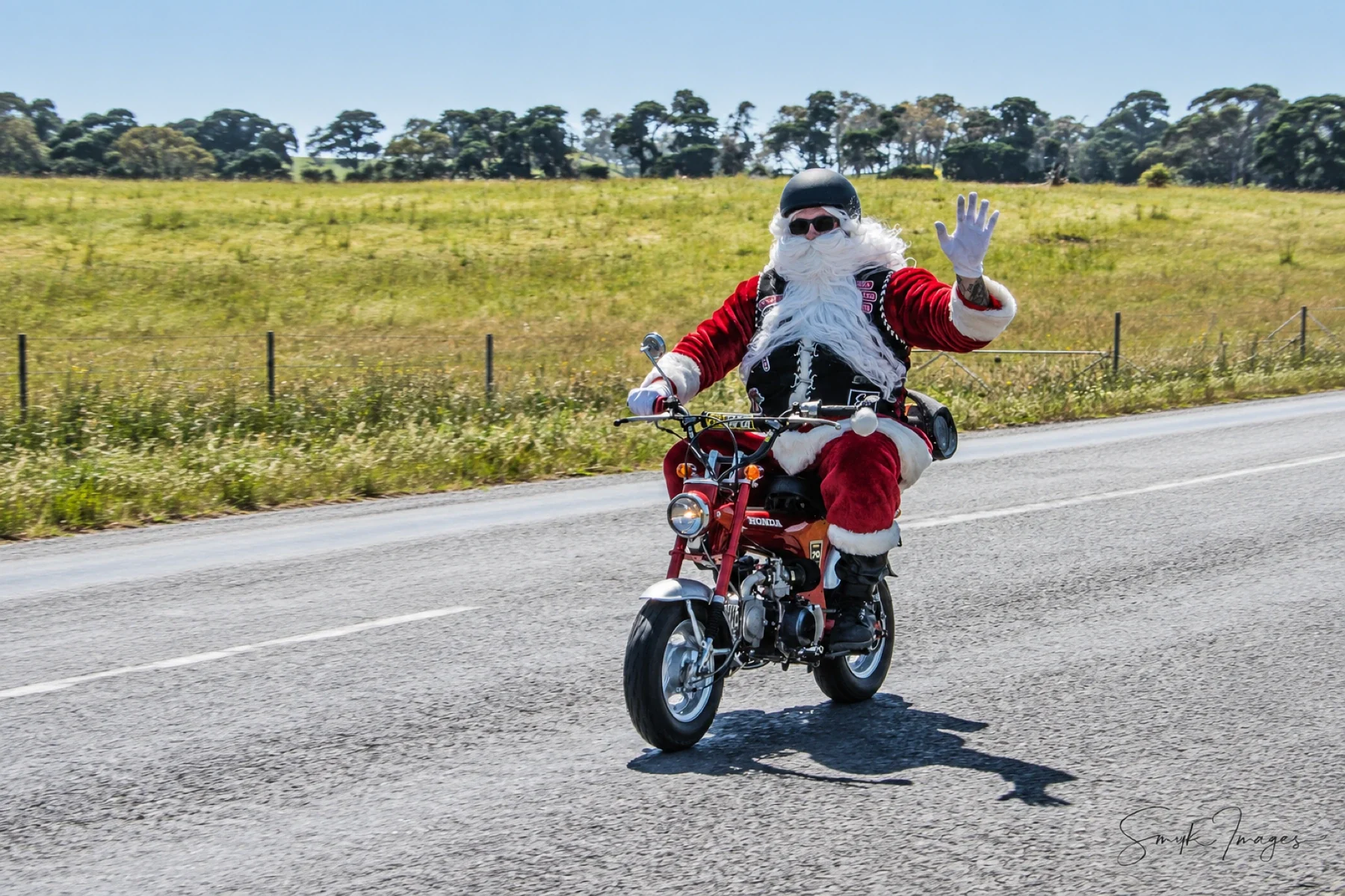 A motorcyclist dressed as Santa waves while riding a small red motorcycle along a rural road beside a grassy paddock.